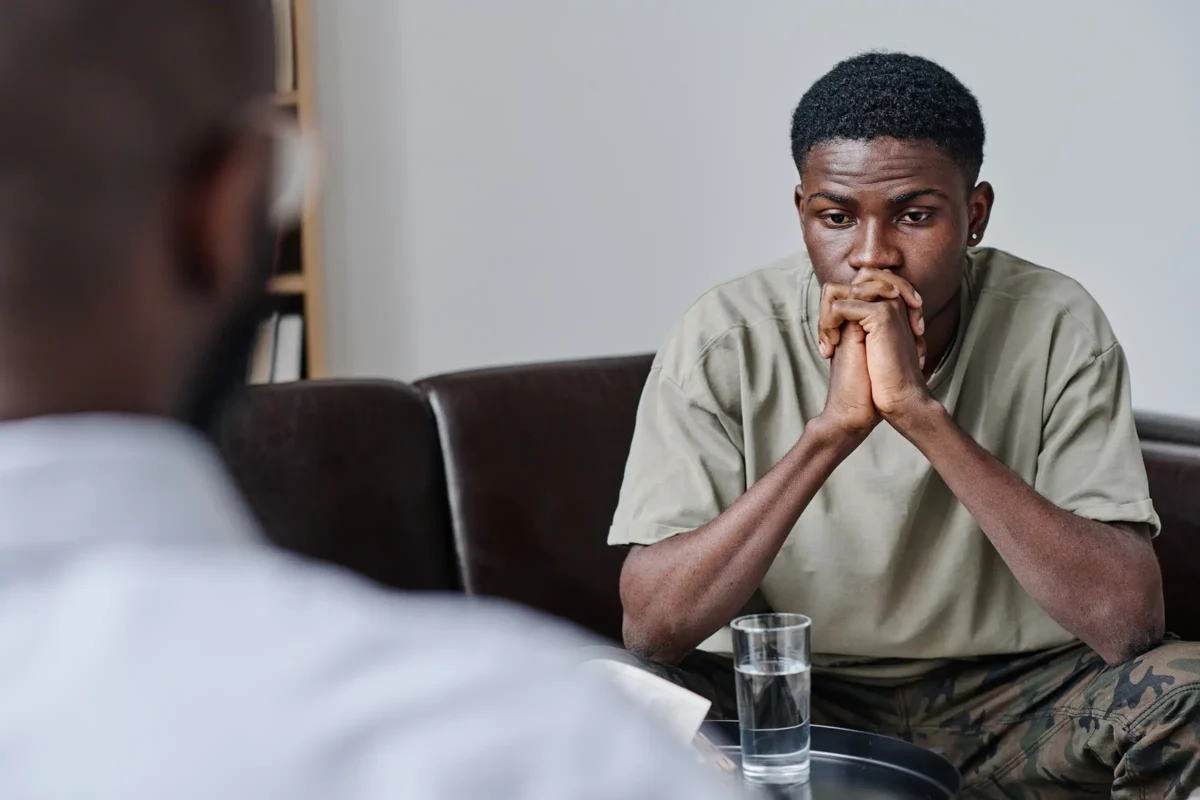 African teenage boy with sad expression sitting at psychologist office and talking to specialist