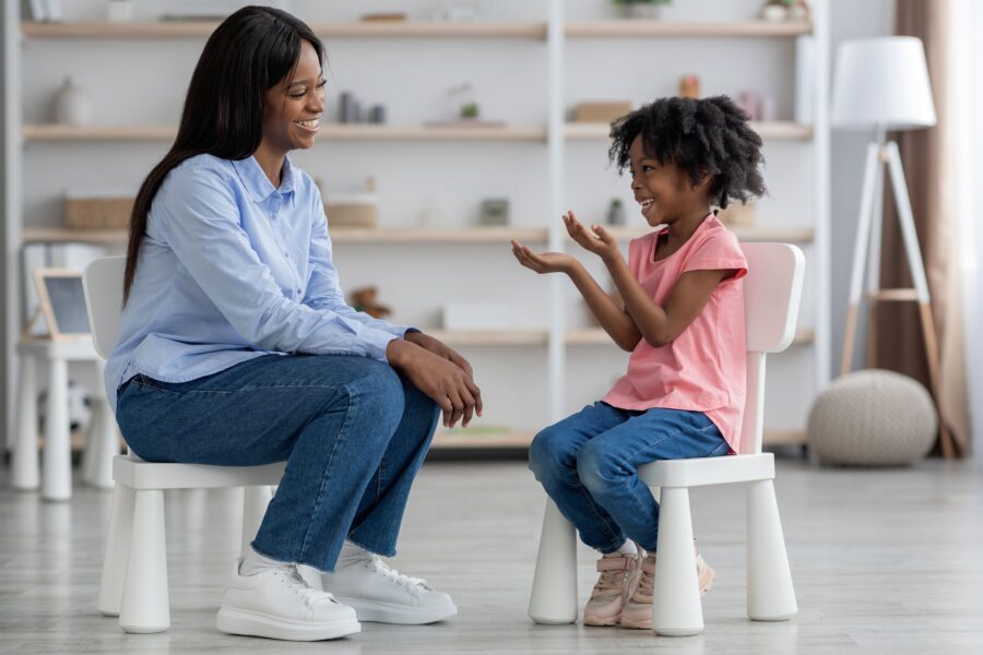 Emotional cute little african american girl at child psychologists office, sitting on chair in front of attractive female therapist, gesturing and smiling, feeling safe. Child psychotherapy concept